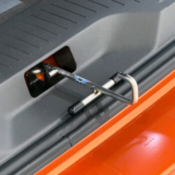 Close-up of a metal U-shaped handle attached to a black rod, mounted on a textured gray vehicle interior panel near an orange edge, likely a cargo area latch or tool attachment.